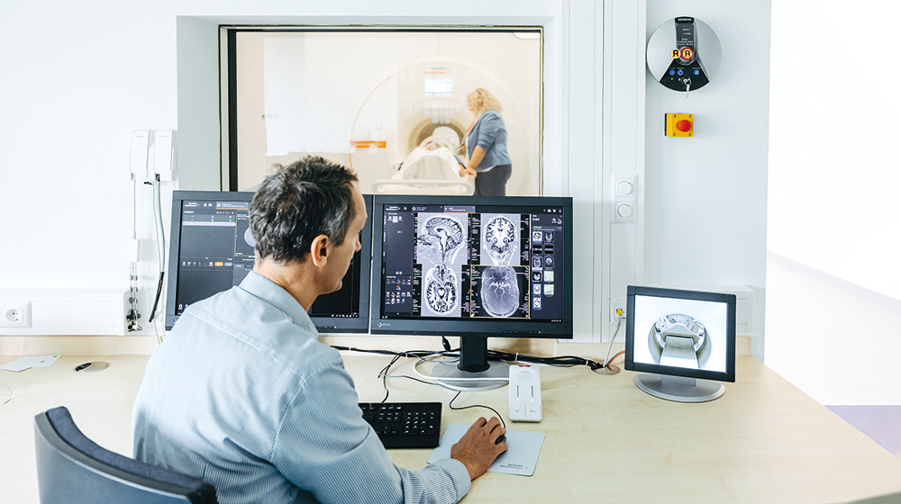 Prof. Speck sits at the workstation of the 7 Tesla MAGNETOM Terra.X Impulse Edition with two screens in front of him with recordings of the brain and watches through a window as a woman operates the MRI (c) Jana Dünnhaupt University of Magdeburg