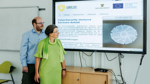 Robert Altschaffel und Jana Dittmann Robert Altschaffel und Jana Dittmann stehen vor einem großen Bildschirm, auf dem ihr Kompass zur digitalen Selbstverteidigung zu sehen ist (c) Jana Dünnhaupt Uni Magdeburg