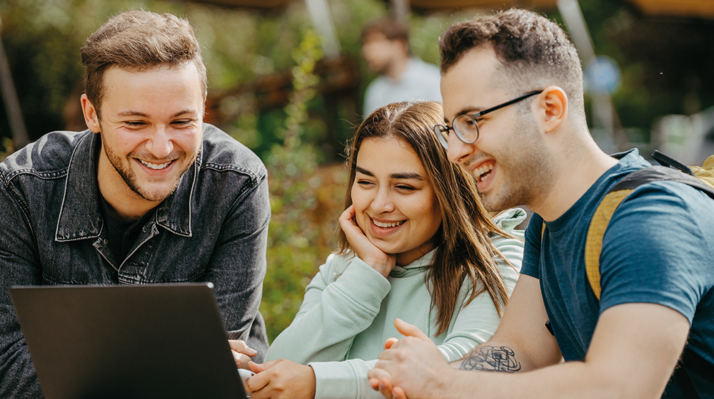 drei Studierende stehen an einem Tisch auf dem Campus und schauen sich gemeinsam etwas auf einem Laptop an (Foto: Jana Dünnhaupt / Uni Magdeburg)